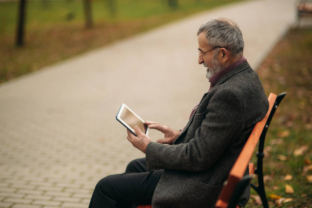 Dziadek korzysta z tabletu i znajduje się w parku na ławce. Patrzył do ekranu - Zdjęcie, obraz