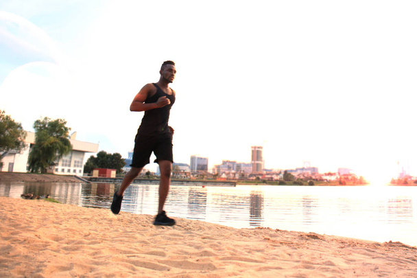 Piena lunghezza di giovane uomo africano in abbigliamento sportivo jogging durante l'esercizio all'aperto, in spiaggia al tramonto o all'alba
. - Foto, immagini