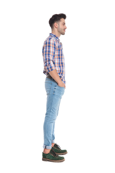 relajado hombre casual con una camisa a cuadros esperando en línea mientras está de pie sobre fondo blanco con las manos en los bolsillos, imagen de longitud completa
 - Foto, Imagen