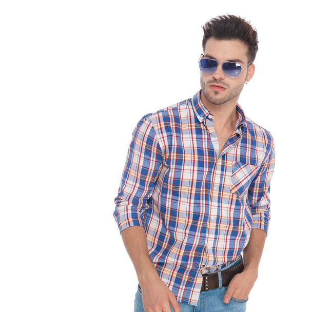 Porträt eines entspannten jungen Mannes mit Sonnenbrille blickt zur Seite, während er auf weißem Hintergrund steht - Foto, Bild