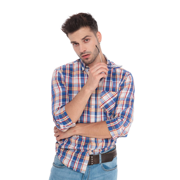 portrait of seductive casual man wearing shirt with blue checkers holding sunglasses while standing on white background - Fotó, kép
