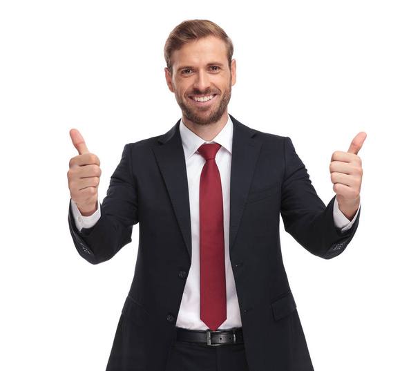 portrait of excited young businessman making thumbs up sign while standing on white background and smiling - Foto, Bild
