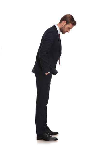 side view of relaxed businessman looking down while standing on white background with hands in pockets, full body picture - Foto, Bild