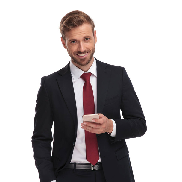 portrait of happy elegant businessman sending an email on his mobile phone while standing on white background with a hand in pocket - Photo, image