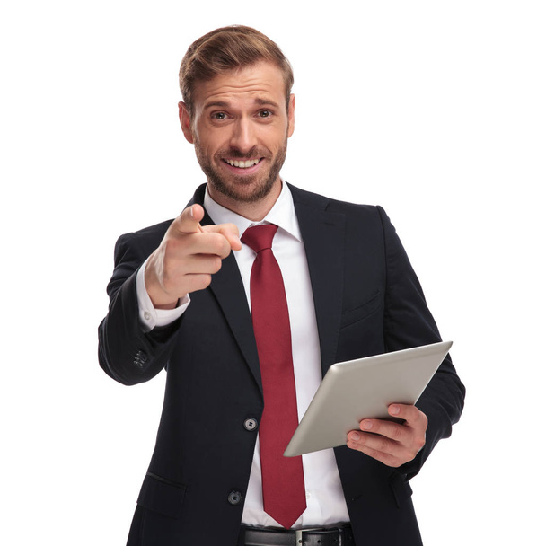 portrait of excited businessman wearing suit and red tie holding tablet and pointing finger while standing on white background - Photo, Image
