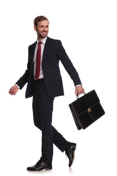 vue de côté de l'homme d'affaires heureux tenant la mallette marchant sur fond blanc et regardant vers le côté, image pleine longueur
 - Photo, image