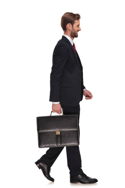 Seitenansicht eines gutaussehenden Geschäftsmannes mit Koffer, der auf weißem Hintergrund läuft und lächelt - Foto, Bild