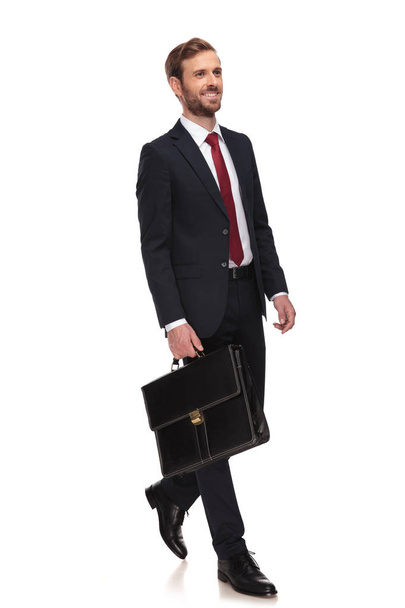 happy young businessman holding briefcase walks to work to side on white background, full body picture - Foto, Imagem
