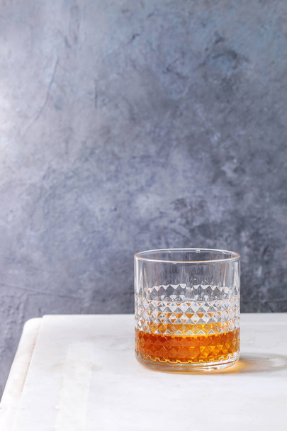 Glass of scotch whiskey standing on white marble table with grey wall at background. Alcohol drink. - Photo, Image