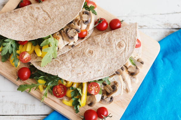 Two whole wheat wraps with chicken breast, mushrooms, yellow bell pepper, cherry tomatoes, arugula and parsley served on wooden board with blue napkin - Photo, Image