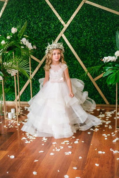 Cute little girl with blond curly hair in a white wedding dress and a wreath of flowers in floral decorations made from tropical leaves, white roses and orchids - Photo, image