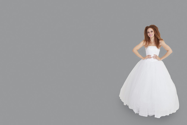Portrait of happy brunette in wedding dress with hands on hips standing over gray background - Photo, image