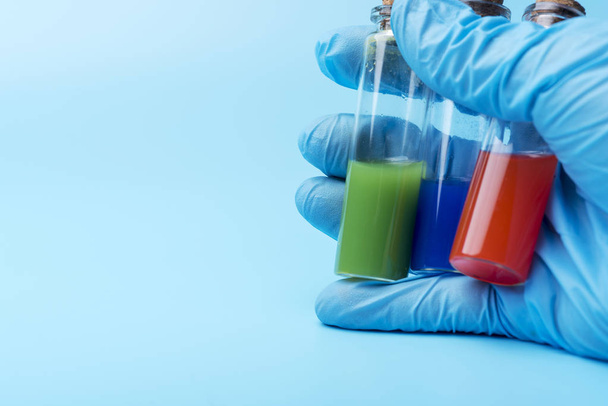 Une main gantée tient trois flacons de liquide différent à l'intérieur sur un fond bleu. Tubes avec liquide rouge, vert et bleu
. - Photo, image