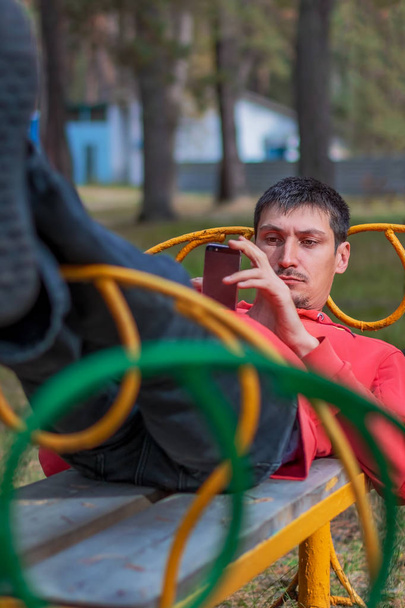 Een blanke man, 30 jaar oud, ligt op een bankje en kijkt naar de telefoon. Hiermee verzendt u berichten en deelt nieuws buitenshuis. - Foto, afbeelding