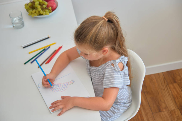 Little girl sitting at home table, absorbedly drawing colorful pictures with pencils - Фото, зображення