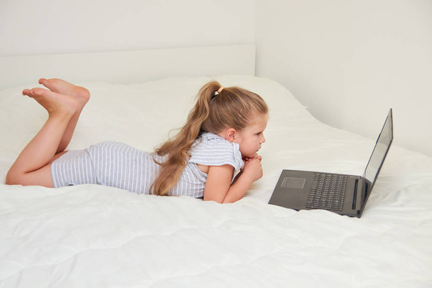 Little blonde girl looking at laptop while lying on bed in her room - Zdjęcie, obraz