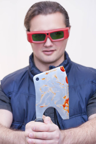 Retrato de hombre joven de etnia caucásica con gafas de sol rojas sosteniendo hacha de cocina cuchilla sangrienta sobre fondo blanco
 - Foto, Imagen