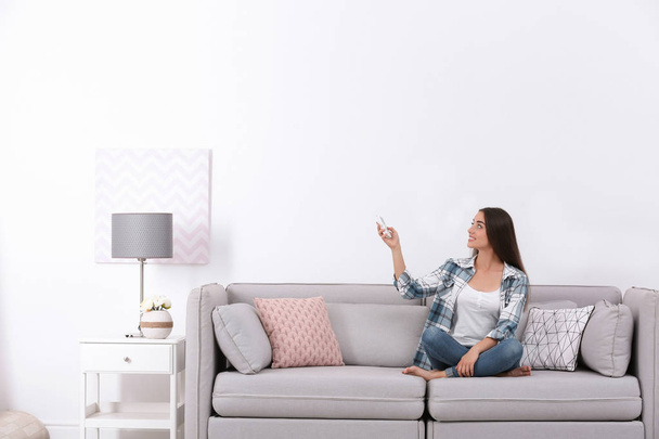 Young woman switching on air conditioner while sitting on sofa near white wall - Foto, Imagem