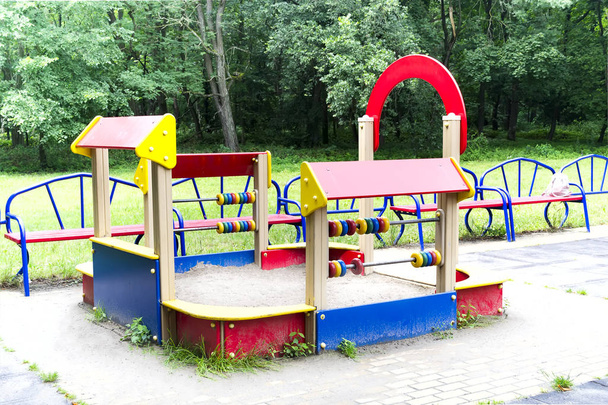Разноцветная детская площадка с песочницей на фоне зеленых деревьев
, - Фото, изображение