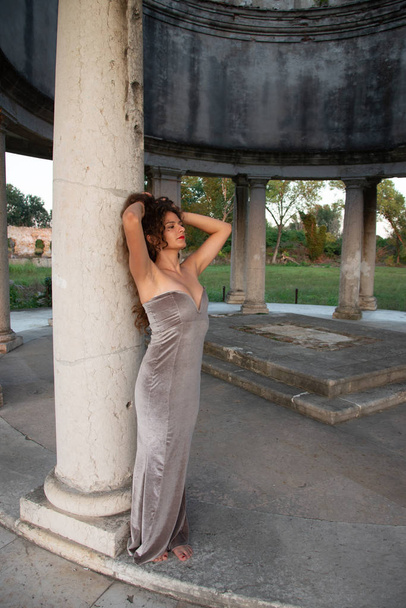 Girl with long wavy brown hair with long elegant dress in anthracite color. Photographed in a classic temple. Full picture. Warm sunlight at sunset. - Photo, Image