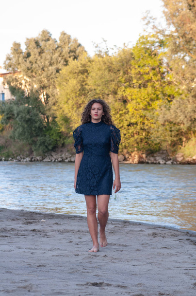 Κορίτσι με καστανά μακριά κυματιστά μαλλιά, περπατάει προσεκτικά κατά μήκος της όχθης του ποταμού. Ζεστό ήλιο στο ηλιοβασίλεμα. Λεπτή λευκή άμμο του κοριτσιού που φοράει ένα φόρεμα με δαντέλα μπλε. - Φωτογραφία, εικόνα