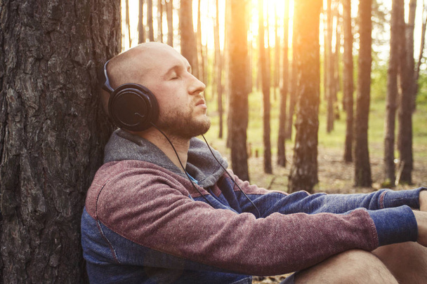 Man in headphones is sitting in a forest with his back to a tree. Concept of outdoor recreation and relaxation. Sounds of nature. - Photo, Image