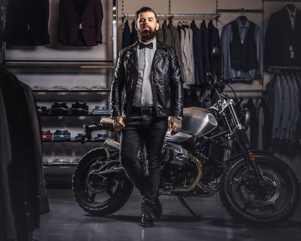 Elegante hombre barbudo tatuado con chaqueta de cuero negro y pajarita posando cerca de la moto deportiva retro en la tienda de ropa para hombre
. - Foto, imagen