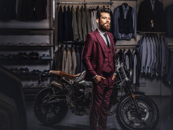 Knappe man met een stijlvolle baard en haren gekleed in vintage rood pak poseren in de buurt van retro sport motor op mens kledingwinkel. - Foto, afbeelding