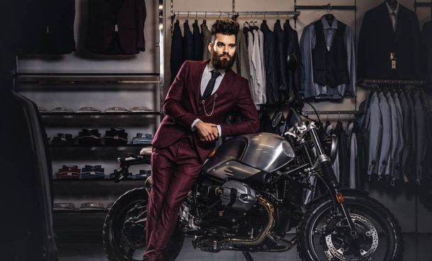 schöner Mann mit stylischem Bart und Haaren im roten Anzug posiert in der Nähe von Retro-Sportmotorrädern im Herrenbekleidungsgeschäft. - Foto, Bild