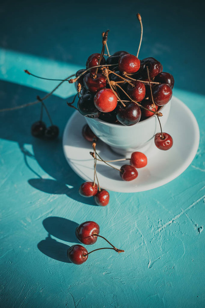 Cherries in the beautiful white cup on turquoise background, fruits, berries.fresh picked cherries. Healthy detox, White cup with ripe berries of red sweet cherries and several fruit - Photo, Image