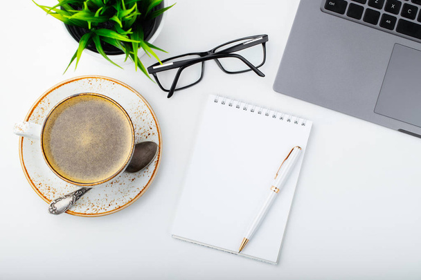 Desk with laptop, eye glasses, notepad, pen and a cup of coffee on a white table. Top view. Flat lay. Light background. - Φωτογραφία, εικόνα