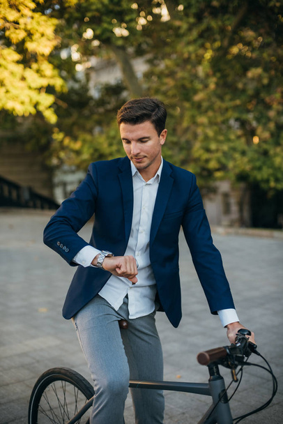 Handsome young man standing with his bicycle on the street in city center and looking on watch - Фото, изображение