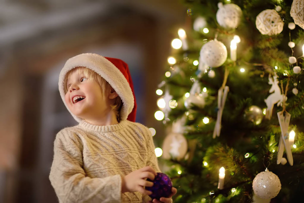 Little boy wearing Santa hat ready for celebrate Christmas. Cute child decorating the Christmas tree with purple glass toy - Φωτογραφία, εικόνα