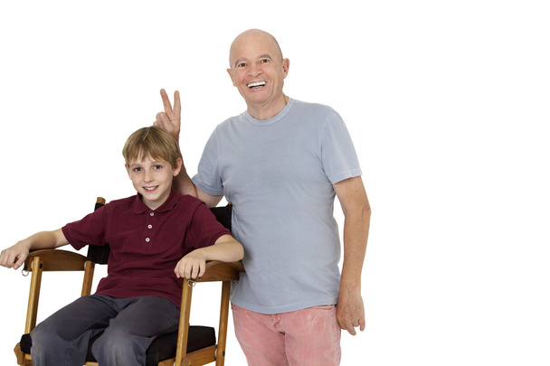 Portrait of senior man gesturing peace sign while pre-teen boy sitting on director's chair over white background - Foto, imagen