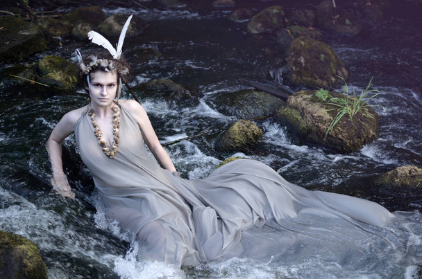 ninfa do rio em um vestido de seda cinza, um colar de conchas de caracol do mar e uma cobertura para a cabeça de perevsidit e descansando em seu rio
 - Foto, Imagem