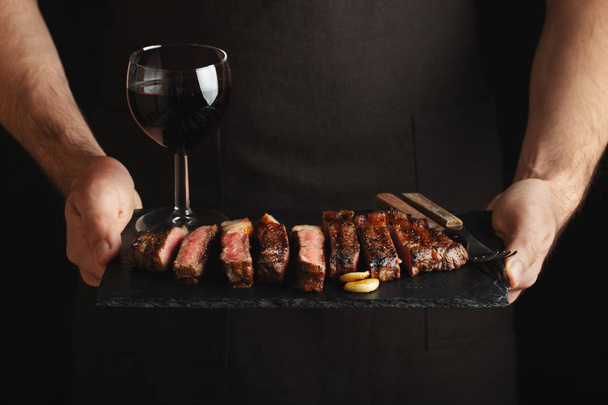 Un homme tenant un steak de bœuf grillé juteux avec des épices et un verre de vin rouge sur une planche à découper en pierre sur fond noir
. - Photo, image