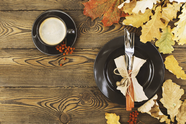 Herbsttisch zum Dankeschön-Essen. eine Tasse Kaffee, ein schwarzer Teller, Besteck, farbige Blätter auf einem Holztisch. das Konzept der Herbstkost. flache Lage. - Foto, Bild