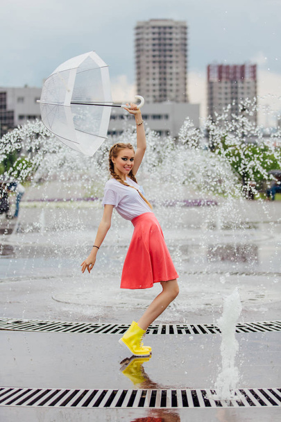 Joven chica bonita con dos trenzas en botas amarillas y con paraguas transparente se encuentra cerca de la fuente. Día lluvioso en la ciudad
. - Foto, Imagen