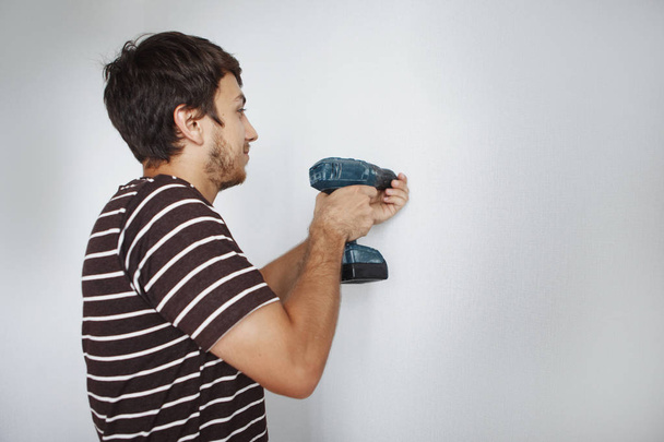Joven chico guapo con un destornillador está perforando una pared blanca en un nuevo apartamento haciendo reparaciones
. - Foto, Imagen