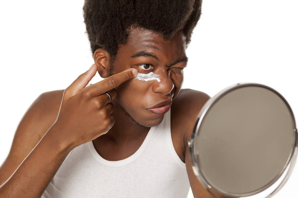 Νέοι Αφροαμερικανός άντρας εφαρμογή κρέμας προσώπου κάτω από τα μάτια του σε λευκό φόντο - Φωτογραφία, εικόνα