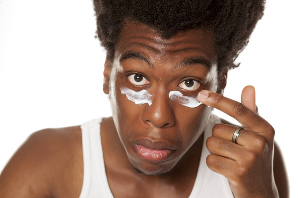 Jonge Afro-Amerikaanse man gezichtscrème onder zijn ogen toe te passen op een witte achtergrond - Foto, afbeelding