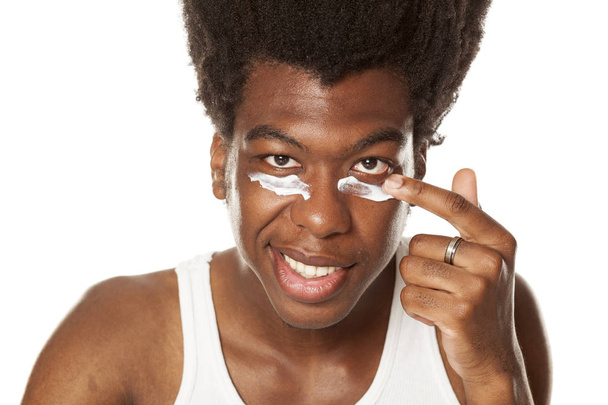 Jeune homme afro-américain appliquant de la crème visage sous ses yeux sur un fond blanc
 - Photo, image