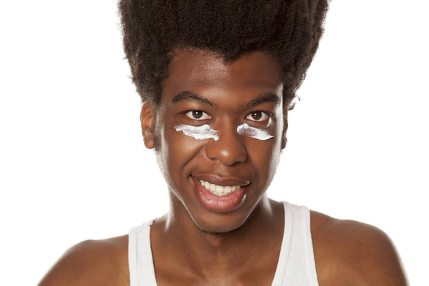 Jonge Afro-Amerikaanse man poseren met gezichtscrème onder zijn ogen op een witte achtergrond - Foto, afbeelding
