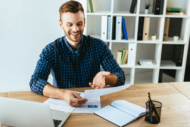 улыбающийся бизнесмен делает бумажную работу за столом с ноутбуком в офисе
 - Фото, изображение