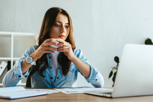 portrait of concentrated businesswoman with cup of coffee at workplace with papers and laptop in office - Фото, изображение