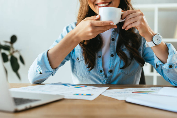 cropped shot of smiling businesswoman with cup of coffee at workplace with papers and laptop in office - Fotografie, Obrázek