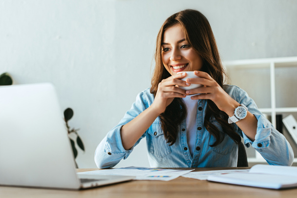 ritratto di donna d'affari sorridente con tazza di caffè sul posto di lavoro con documenti e laptop in ufficio
 - Foto, immagini
