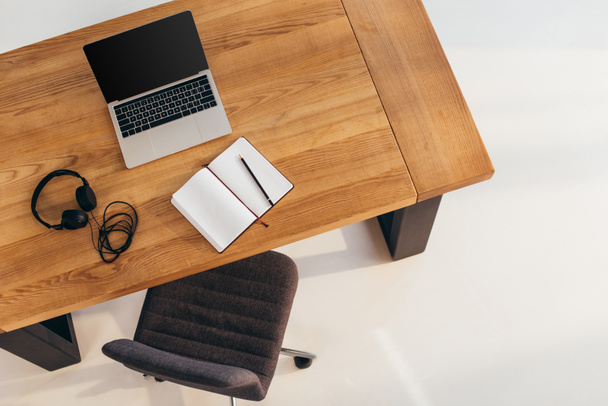vista superior do portátil com tela em branco, fones de ouvido e notebook na mesa de madeira com cadeira de escritório perto por
 - Foto, Imagem