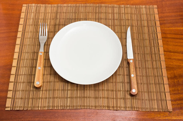 Empty round white dish and cutlery set consisting of the fork and knife with wooden handles on bamboo table mat on a dark red wooden table - Photo, Image