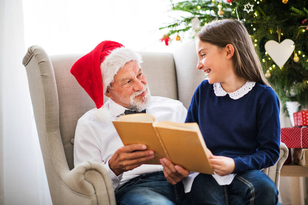 A small girl and her grandfather with Santa hat reading a book at Christmas time. - Foto, Imagem